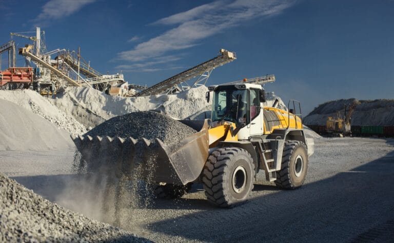 Yellow front-end loader dumping gravel at an open-pit quarry with conveyor belts and large rock piles against a blue sky